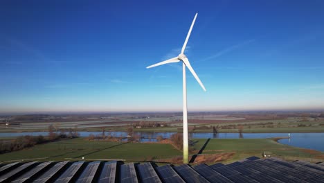 Frozen-solar-panels-in-front-of-solitary-wind-mill-turbine-part-of-sustainable-industry-in-Dutch-flat-river-landscape-against-blue-sky