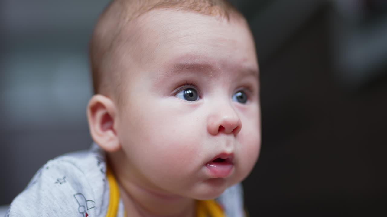 Beautiful face of a small Caucasian infant boy lying on his belly. Cute baby looking around with surprise. Close up. Blurred backdrop.