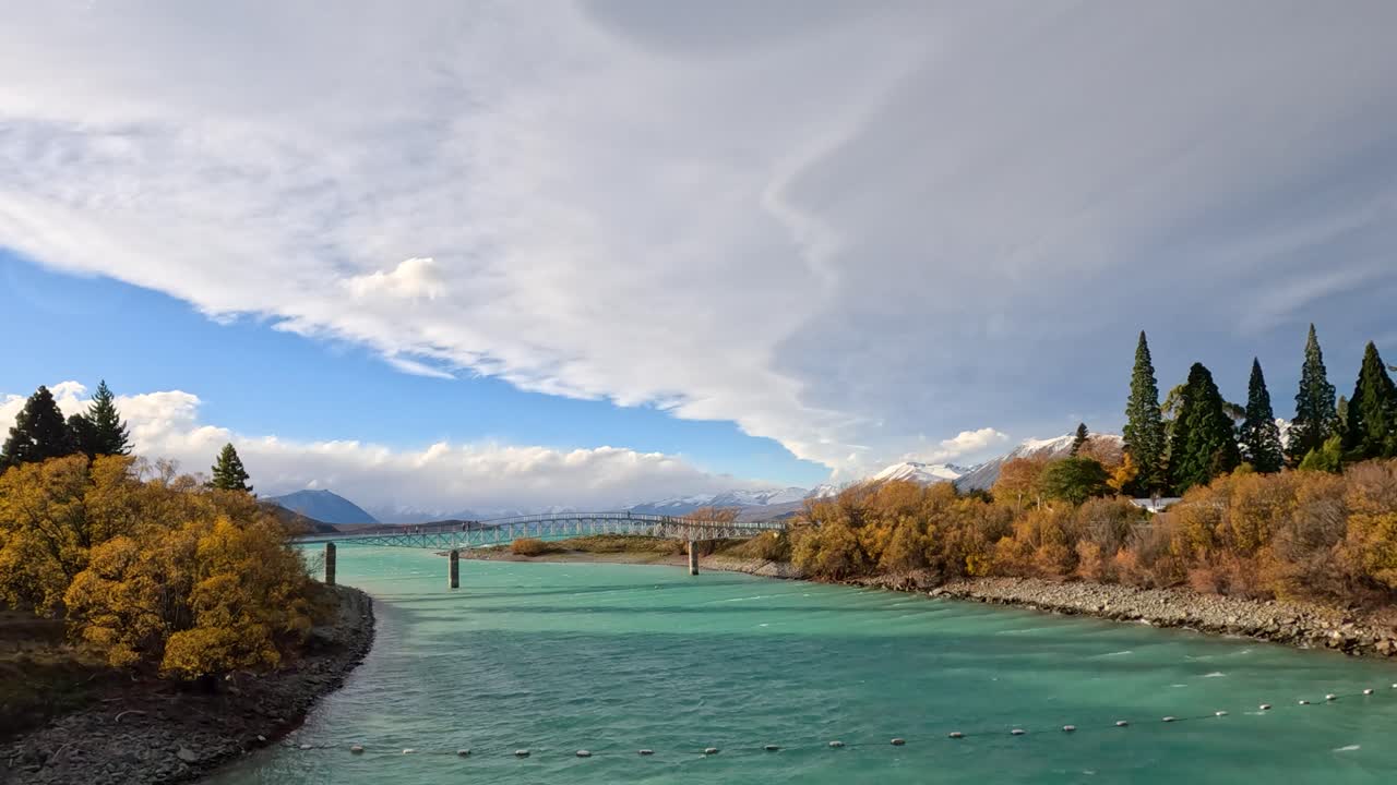 Vehicle travels over a bridge spanning Lake Tekapo, framed by autumn foliage, turquoise water, dramatic clouds, and distant mountains. Smooth forward camera motion, natural daylight