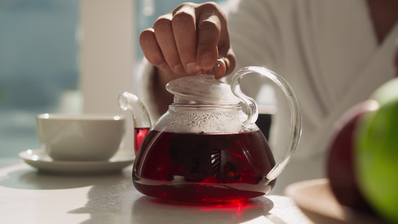 Man closes teapot with tea using lid at table. Final step of brewing hibiscus tea to get natural flavor from tea-time. Healthy homemade beverage