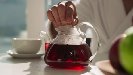 hombre cierra la tetera con té usando la tapa en la mesa. último paso de la elaboración de té de hibisco para obtener el sabor natural de la hora del té. bebida casera saludable