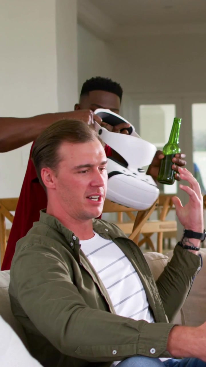 Pystyvideo: Mies näkee pullon VR-laseissa, nojaa sohvan yli ja poistaa lasit puhdistaakseen ne.