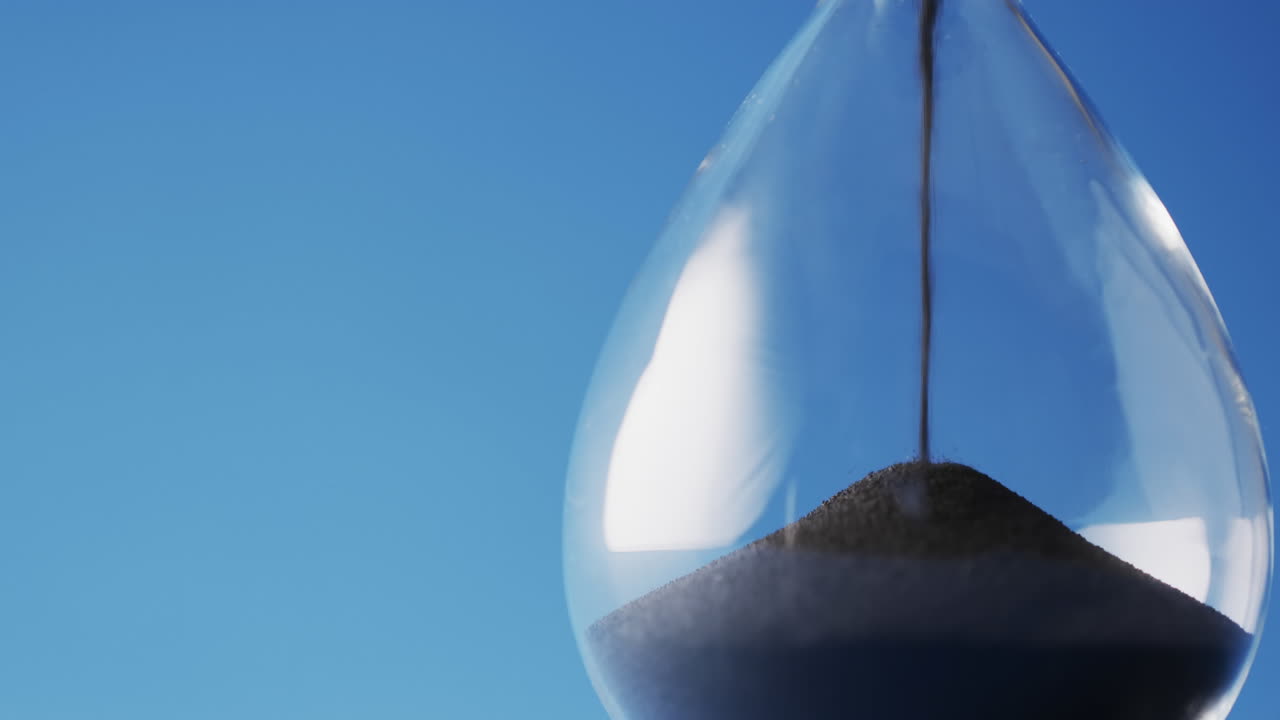 Video of close up of hourglass with black sand pouring with copy space on blue background