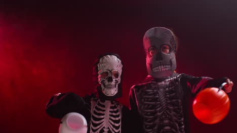 studio shot of children dressed up in skeleton costumes trick or treating with candy buckets at halloween against red smoke background lighting