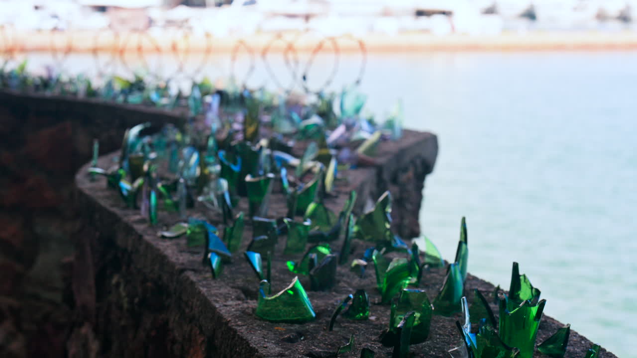 Close up of multiple broken glass shards and barbed wire loops along the edge of a brick wall with a blurred view of the sea