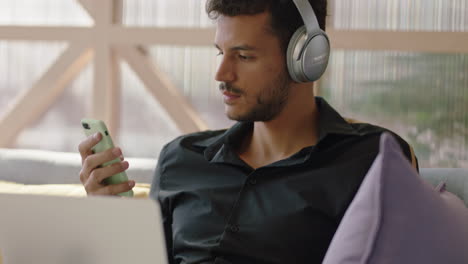 young-hispanic-man-using-laptop-computer-browsing-online-messages-sharing-network-communication-student-enjoying-listening-to-music-checking-smartphone-social-media-in-modern-office