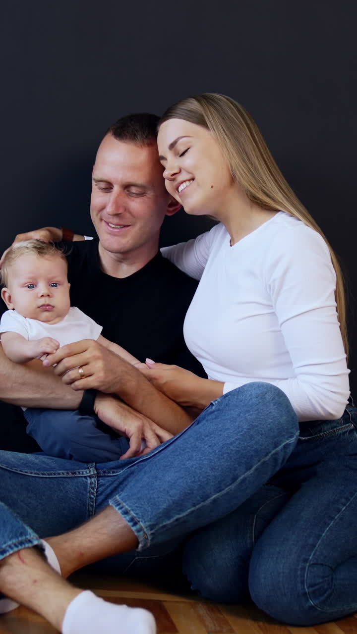 Young modern family wearing jeans sit on the floor with baby in hands. Happy couple kissing. Black backdrop. Vertical video