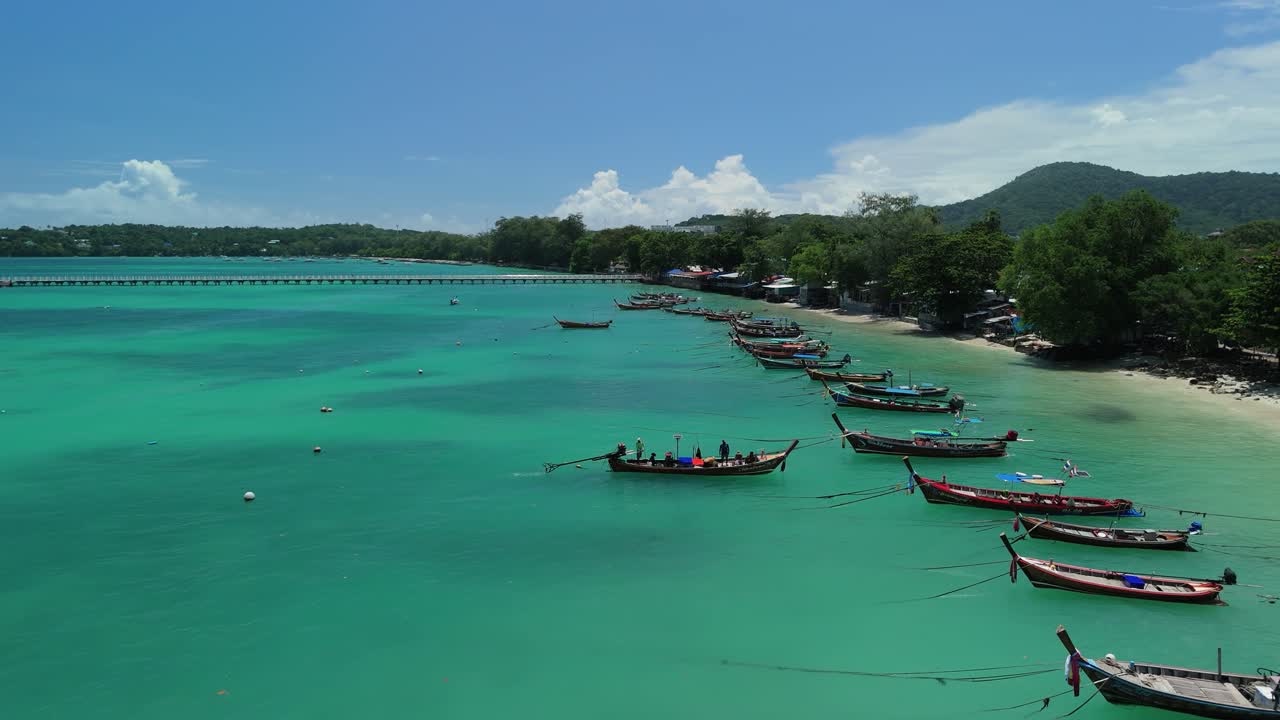 Drone orbits a longtail boat approaching Rawai Beach over turquoise shallows of Phuket