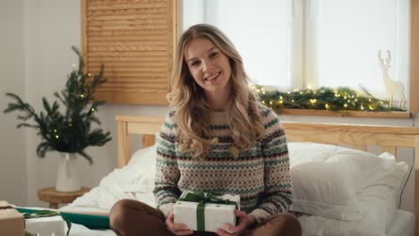 Portrait-of-cheerful-caucasian-woman-holding-a-Christmas-gift-while-sitting-on-bed