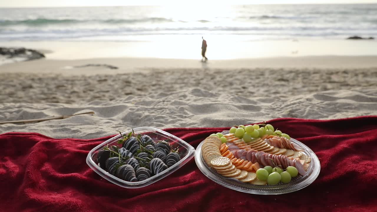 저녁에 두 사람이 멀리 걸어가는 모래 해변에서 빨간 담요에 담긴 과일과 고기의 작은 소풍