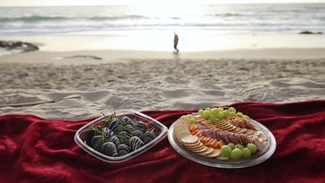 A-small-picnic-of-fruit-and-meat-served-on-a-red-blanket,-on-a-sandy-beach-as-two-people-walk-by-in-the-distance-in-the-evening