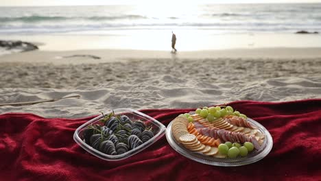 Ein-Kleines-Picknick-Mit-Obst-Und-Fleisch,-Serviert-Auf-Einer-Roten-Decke,-An-Einem-Sandstrand,-Während-Abends-Zwei-Menschen-In-Der-Ferne-Vorbeigehen