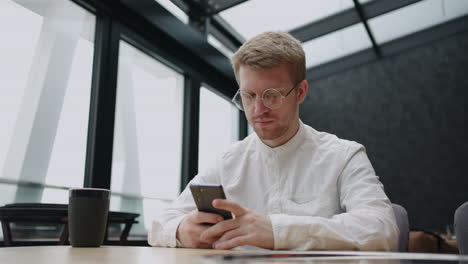 Un-Joven-Con-Gafas-Y-Un-Teléfono-Móvil-En-Las-Manos-Mira-La-Pantalla-E-Imprime-Un-Mensaje.-Hipster-Con-Gafas-Redondas-Escribe-Un-Mensaje-En-Su-Teléfono