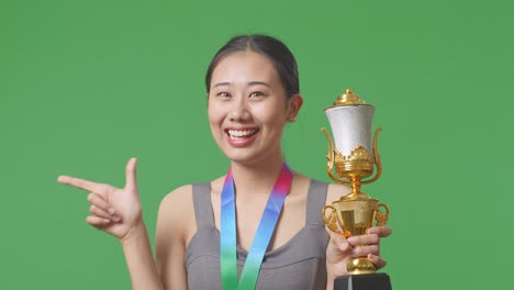 close up of asian woman with a gold medal holding a gold trophy in her hands, smiling, and pointing to side on green screen background in the studio