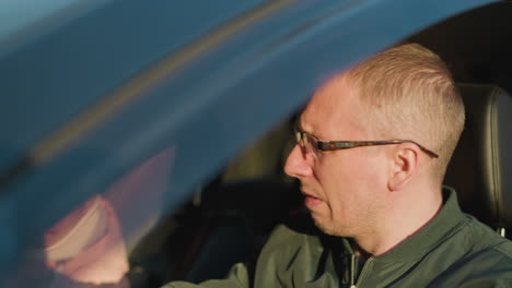 a man wearing a green jacket sits inside a car, carefully adjusting his glasses. in the background, a little girl in a pink jacket and knit hat is also visible inside the vehicle