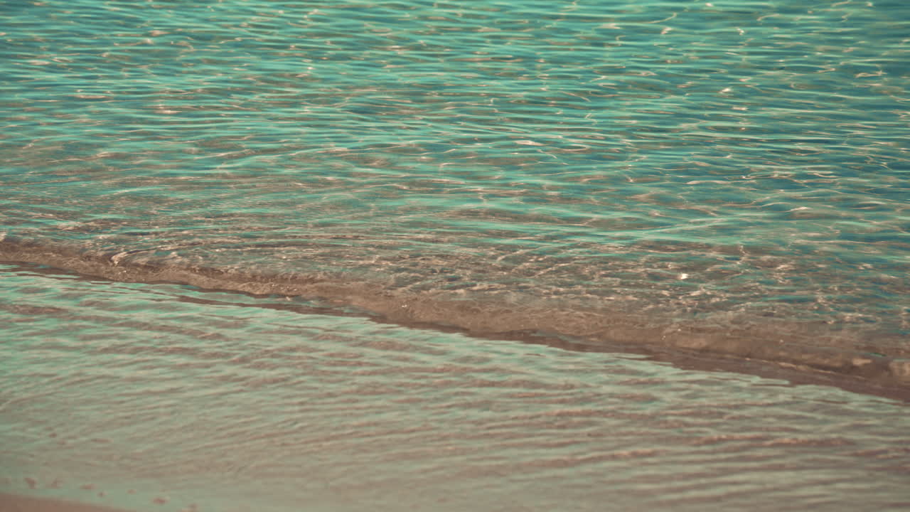 Close up of clear turquoise water gently washing over the sandy shoreline, with soft ripples and a calm, tropical feel