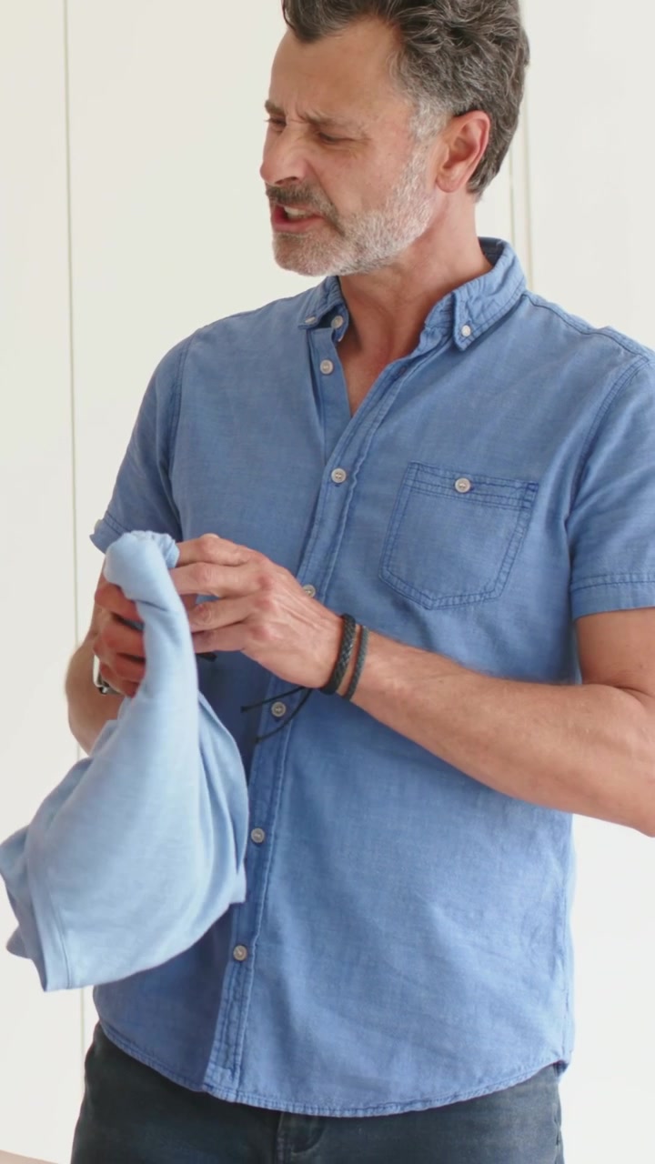 Video verticale: Raggiungendo una scatola aperta, un uomo controlla e piega una camicia azzurra in camera da letto per fare i bagagli.