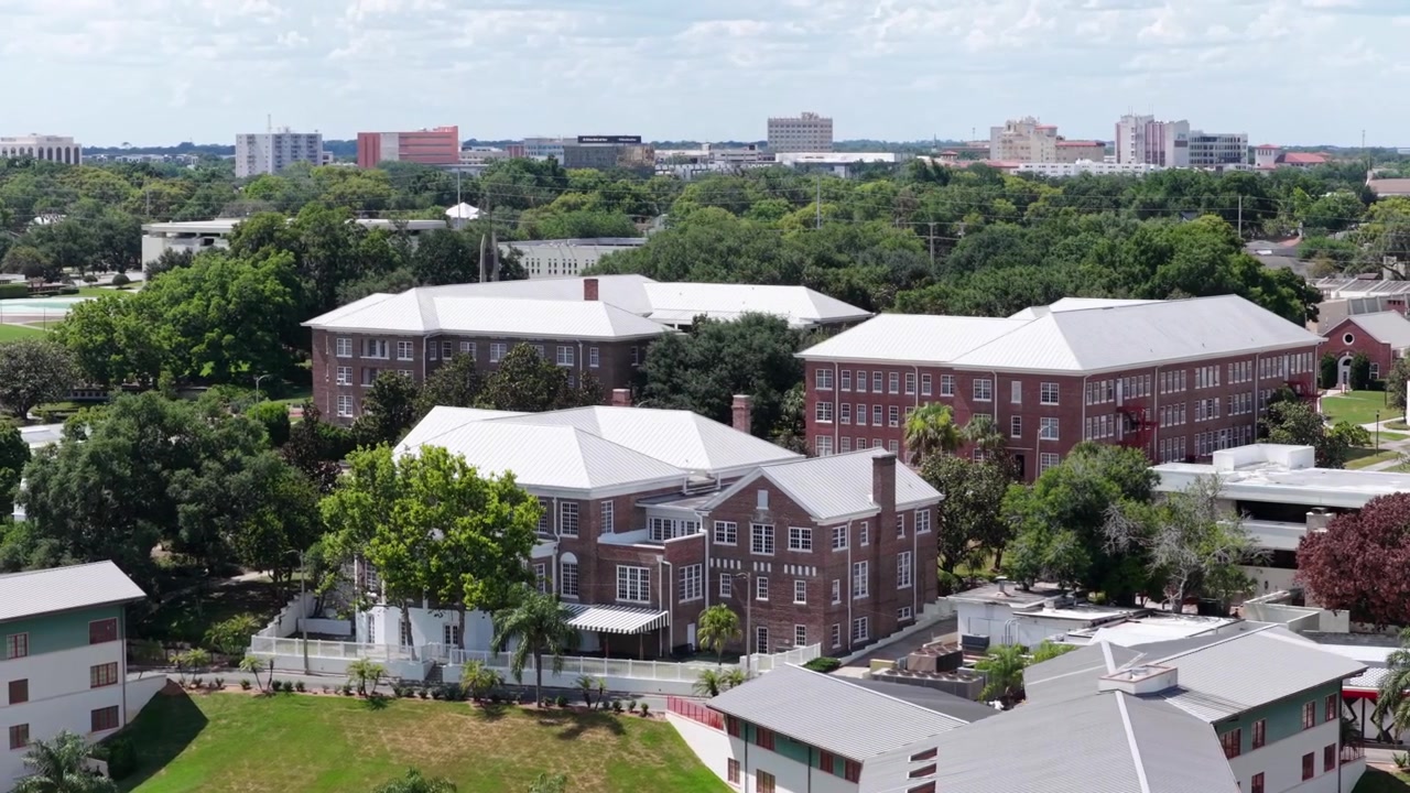 Storici edifici dormitorio in mattoni rossi sono il fulcro del campus verdeggiante del Florida Southern College, rigogliose palme e prati verdi si estendono verso lo skyline del centro di Lakeland in lontananza. Ripresa aerea con drone in orbita.