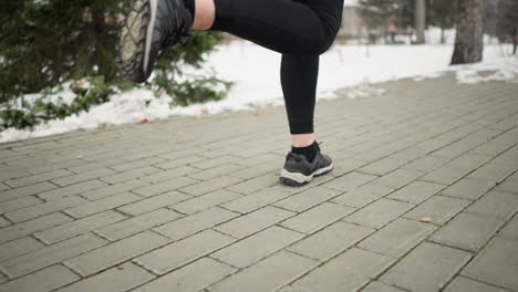 close-up leg view of a lady jogging outdoors during winter on a paved pathway with blurred snow-covered trees and offices in the background, showcasing movement