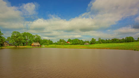 Filmische-Zeitrafferaufnahme-Von-Wolken,-Die-Sich-Schnell-Am-Himmel-über-Einem-See-Mit-Gras-Und-Einem-Haus-Im-Hintergrund-Bewegen,-Zeitraffer