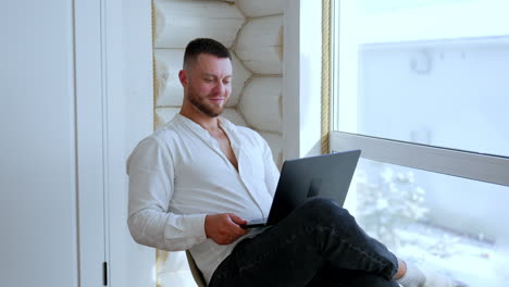 positive relaxed bearded male sits on the chair near the window. man works on laptop from home.