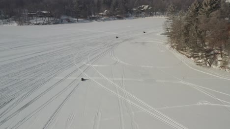 Aerial,-three-people-riding-snowmobiles-on-frozen-lake-covered-with-thick-snow