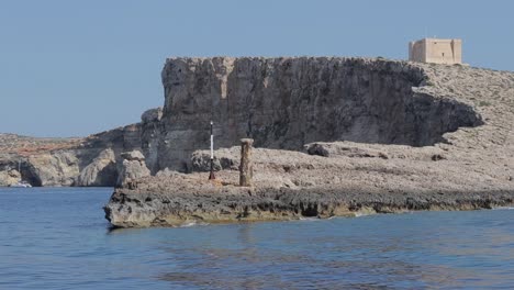 Steile-Klippen-Der-Insel-Comino,-Malta,-Mit-Dem-Turm-Der-Heiligen-Maria-Mit-Blick-Auf-Das-Glitzernde-Meer-An-Einem-Sonnigen-Sommertag