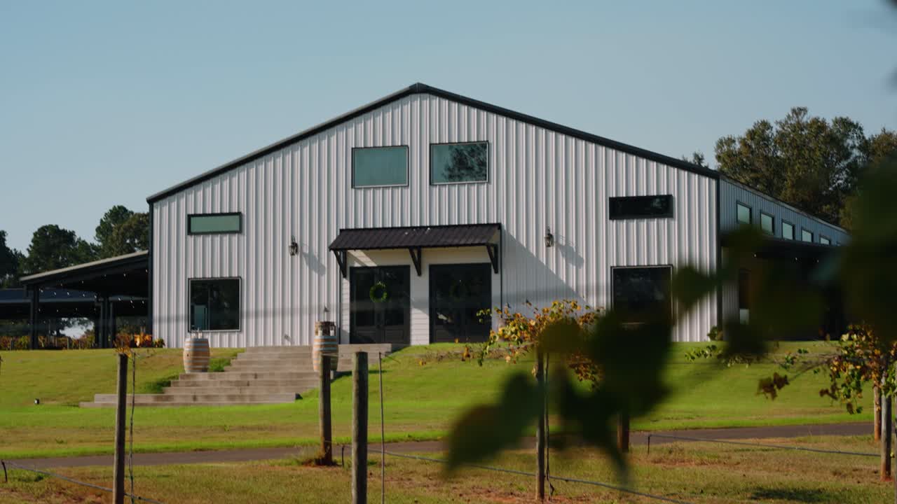 A reveal shot of a countryside wedding venue at a South Georgia vineyard, framed through vibrant leaves. The white farmhouse-style building stands beautifully against a clear blue sky