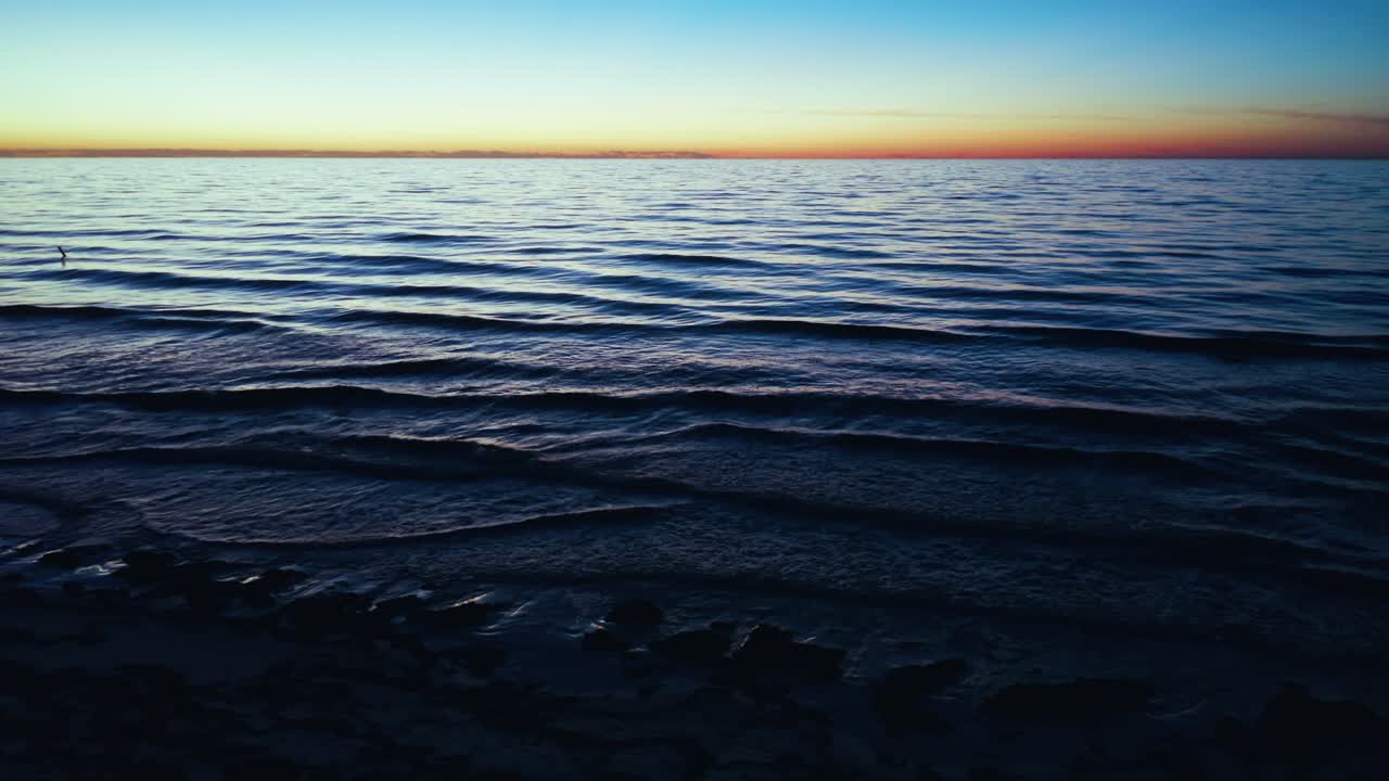 Calm ocean waves flow over a rocky shore as dusk settles, displaying serene water textures and a colorful sky that fades into twilight along the horizon