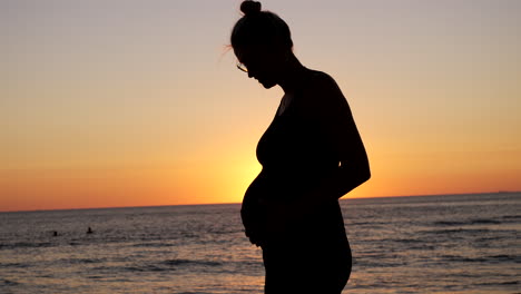 silhouette of pregnant woman touching belly in front of golden sunset at ocean - slow motion close up