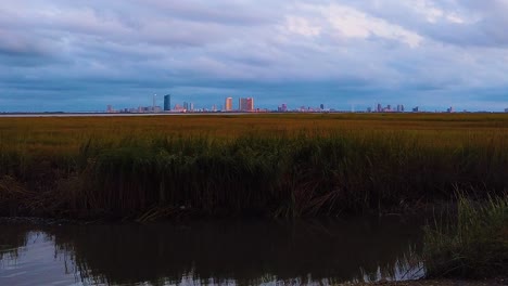 HD-MOTIONLAPSE-Straight-tilt-up-Atlantic-City-skyline-in-distance-over-waterway-with-mostly-cloudy-sky-day-to-night-with-clouds-turning-pink,-purple-and-blue