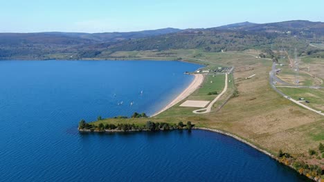 Seestrand-Mit-Menschen-Zum-Sonnenbaden-Und-Baden,-Kleine-Segelboote-Zum-Segeln,-Garten-Und-Spazierbereich-Mit-Den-Häusern-Im-Hintergrund