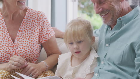 Cerca-De-Abuelos-Sentados-En-El-Sofá-Con-Su-Nieta-En-Casa-Leyendo-Un-Libro-Juntos