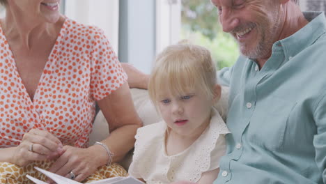 close up of grandparents sitting on sofa with granddaughter at home reading book together