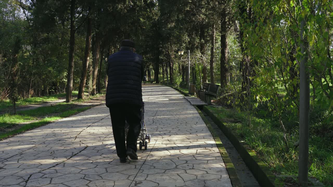 노인은 푸른 나무로 둘러싸인 조용하고 화창한 가을날 이른 아침 공원 도로에서 유모차를 밀고 있습니다.