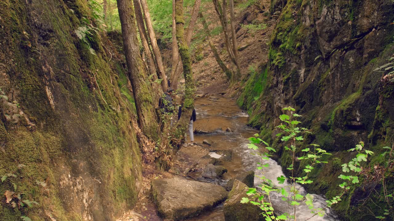 Trekkers walking along stone path near river in forest on mountain slope in sunny weather in summer. Hiker climbs down A Rocky Mountain With Narrow Path, hiking concept. Shot in 4k.
