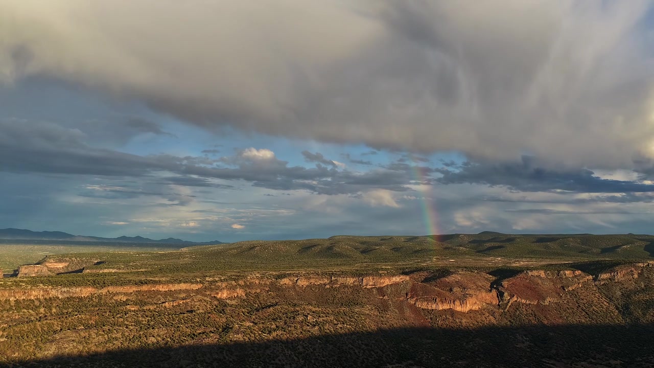 Timelapse 4K UHD con drone che cattura un arcobaleno che svanisce toccando terra in un lussureggiante canyon del sud-ovest. Nuvole temporalesche scure passano sopra mentre la calda luce del tramonto e le ombre profonde attraversano il terreno verde e accidentato.