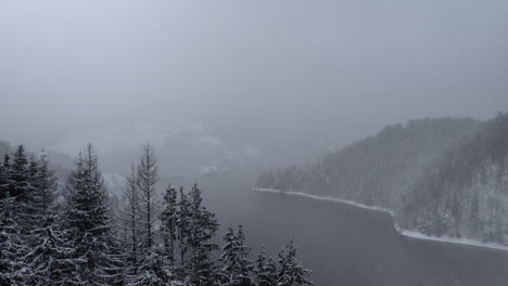 Aerial-panorama-of-a-lake-and-dam-surrounded-by-a-captivating-blend-of-fog-and-snow,-mountain-forest,-aerial-lifting-up,in-Dragan-dam-from-Transylvania,-Romania