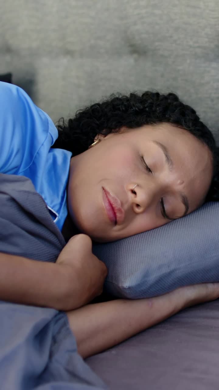 Video vertikal: Kelopak mata berkedip, wanita berkaos biru terbangun berbalik ke kanan di tempat tidur memegang selimut