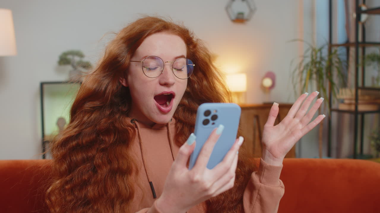 Surprised caucasian redhead woman with windswept hair using smartphone sitting on sofa at home