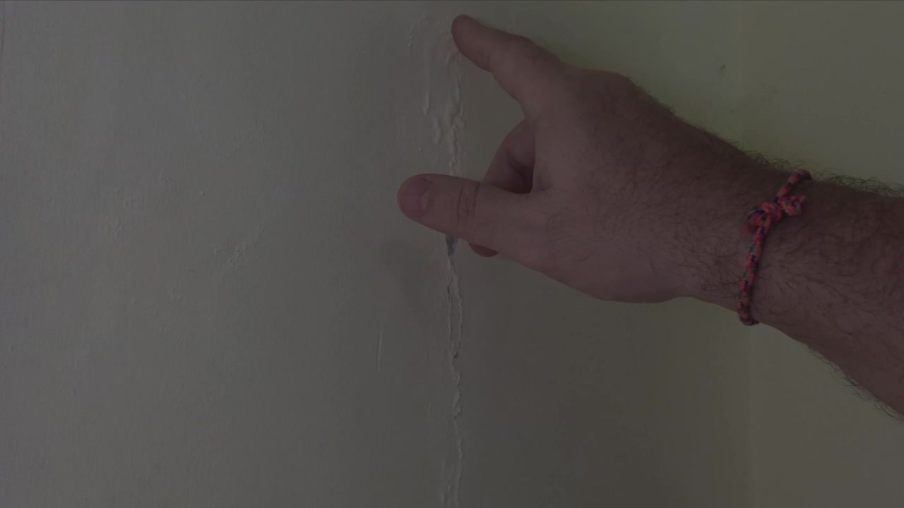 Primo piano di un dito indice che punta direttamente a una crepa verticale con vernice che bolle e si scrosta su una parete interna durante un'ispezione per danni domestici.
