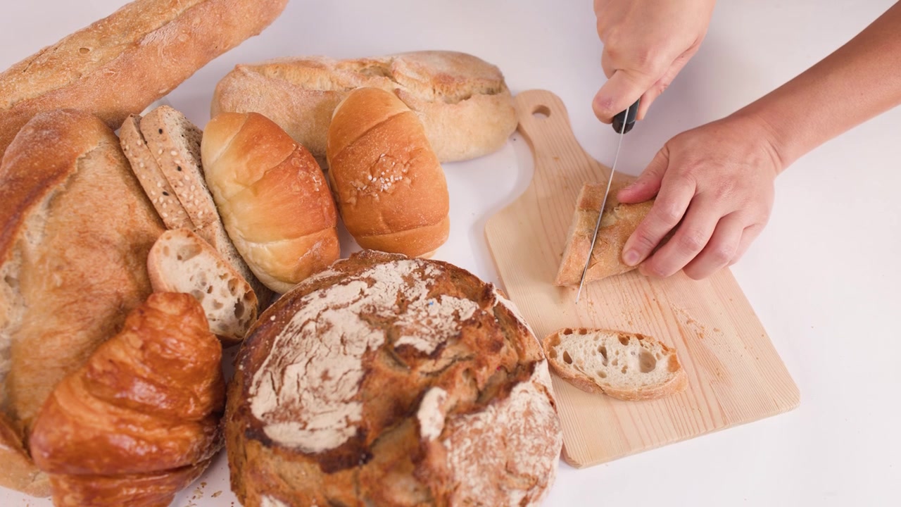 Close-up of hands cutting bread on a wooden board surrounded by various artisan pastries.