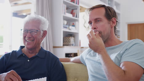 father with adult son eating popcorn watching movie on sofa at home together