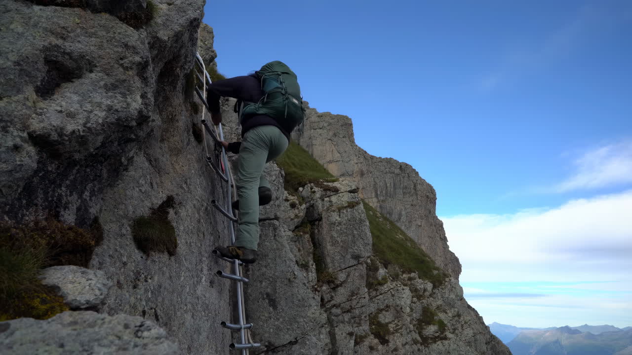 Alpinista Caucasiano Subindo Escada de Ferro em Cume Alpino Exposto, Usando Mochila e Arnês, Agarrando Metal