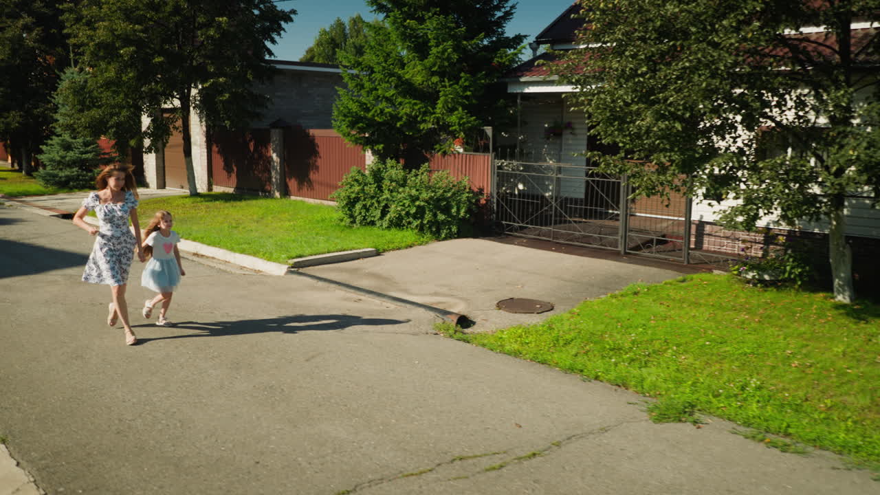 Young woman in floral dress runs hand in hand with little cousin on quiet sunny street, both smiling as their hair sways with motion, surrounded by green lawn, gated homes, and tall trees