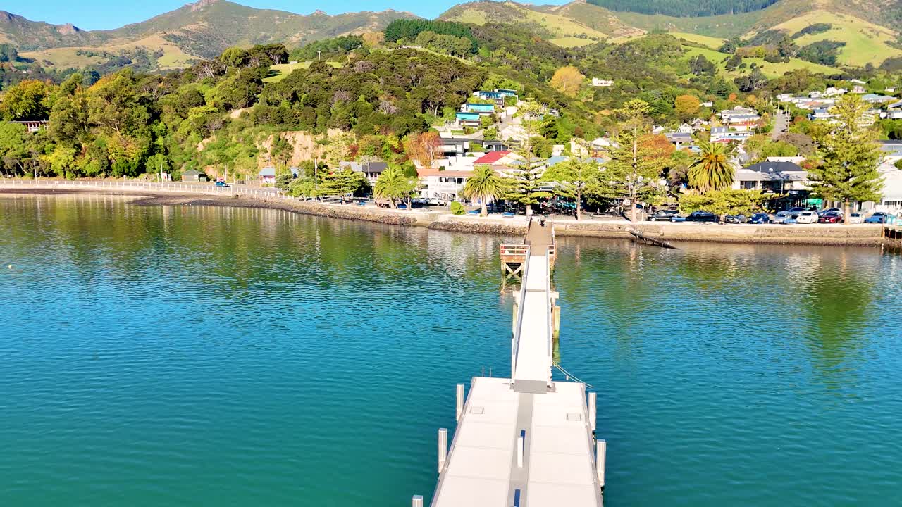 Aerial footage of Akaroa pier with vibrant hillside homes, captured in bright daylight, showcasing serene waters and lush greenery