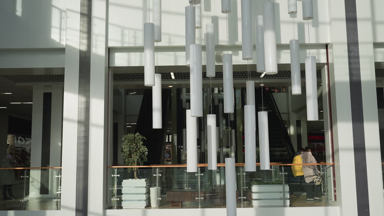 Modern shopping mall interior featuring suspended white tube lights, escalator descending between glass railings, decorative plants, and people walking through brightly lit commercial space