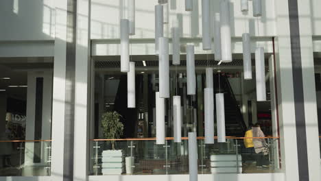modern shopping mall interior featuring suspended white tube lights, escalator descending between glass railings, decorative plants, and people walking through brightly lit commercial space