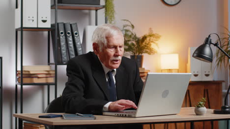 bored sleepy senior businessman working on laptop computer yawns leaning on hand at office desk