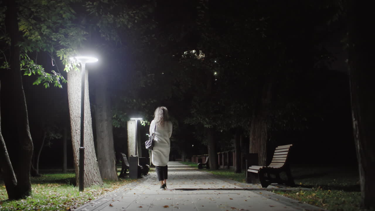 Back view of girl with curly hair in white coat carrying black handbag, walking alone down empty park walkway at night, surrounded by benches, trees, fallen leaves and illuminated by streetlamps
